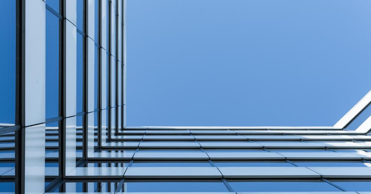 „Clădire modernă cu fațadă din sticlă, fotografiată de la nivelul solului în unghi ascendent, cu linii geometrice simetrice și reflexii ale cerului albastru pe panourile de sticlă.